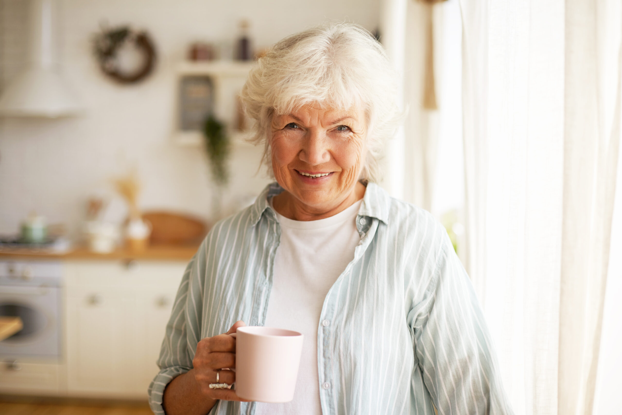 Positive friendy looking senior elderly woman with gray hair and wrinkles spending day at home, posing by window with cozy clean kitchen in background, drinking tea or coffee in the morning support at home classifications 3 and 4