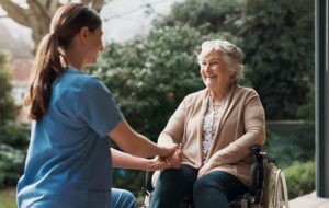 A nurse in blue scrubs holds the hand of an elderly woman in a wheelchair, showing support available for older Australians eligible for Support at Home