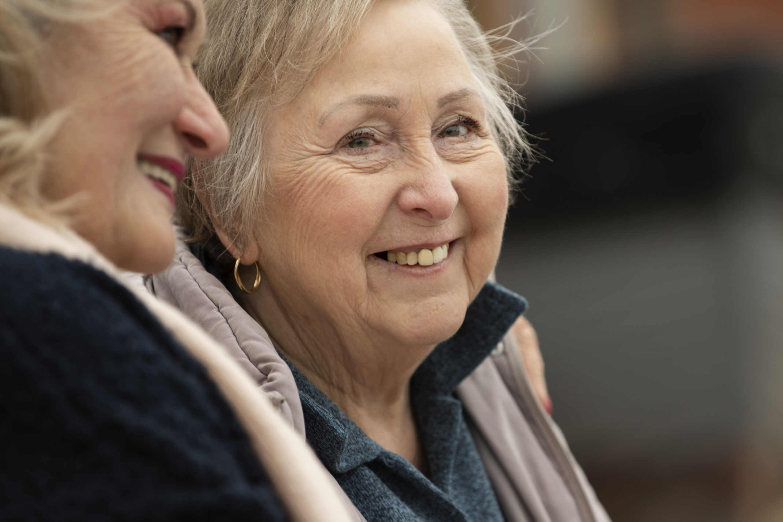 happy-senior-women-friends-chatting Two older women smiling and embracing, representing the security and support that grandfathered pensioners retain under the Support at Home program.